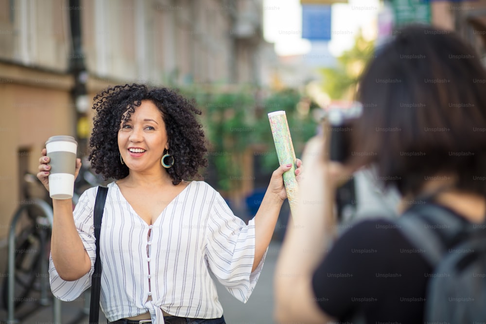 Smiling woman standing on the street with cup of coffee. Woman taking photo of her friend. Focus is on background.