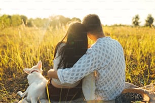 Stylish couple embracing in sunny light, relaxing with white dog on blanket among grass in summer meadow. Summer vacation and picnic. Young family enjoying sunset with swiss shepherd puppy