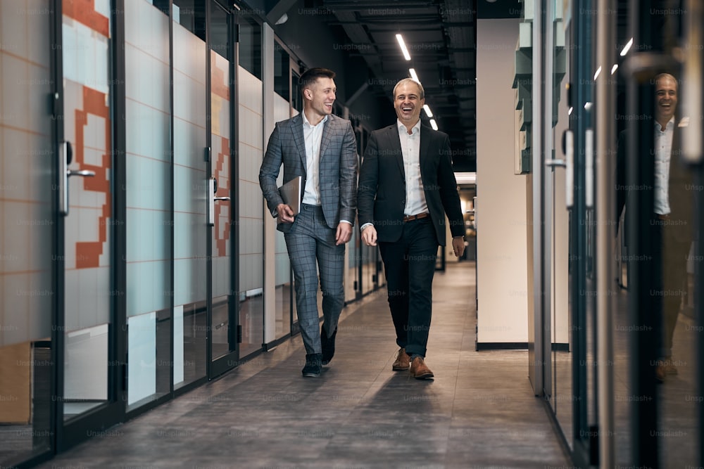 ハンサムなビジネスマンは、ラップトップを保持し、同僚とオフィスの廊下を散歩しながら微笑む