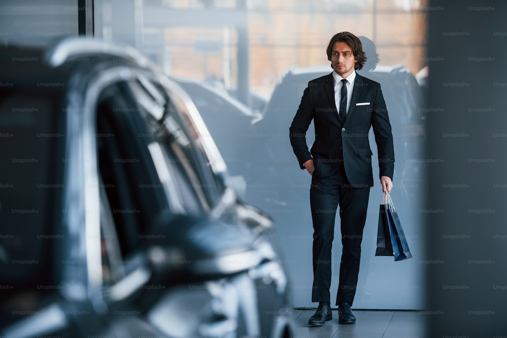Retrato de un joven y guapo hombre de negocios con traje negro y corbata y con bolsa de compras cerca del automóvil moderno.