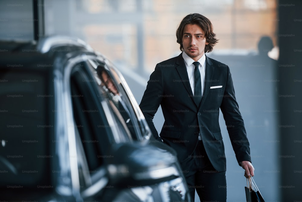 Retrato de um belo jovem empresário de terno preto e gravata e com sacola de compras perto do carro moderno.