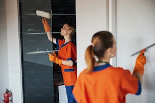 Joyous beautiful Caucasian female cleaner in the uniform and her colleague washing the glass walls
