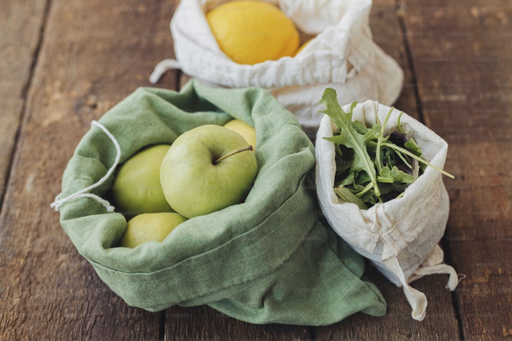 Compras de comestibles sin plástico. Manzanas frescas, limones y rúcula en bolsas de algodón ecológico sobre mesa rústica de madera. Residuo cero. Frutas y verduras ecológicas en bolsas reutilizables. Estilo de vida sostenible