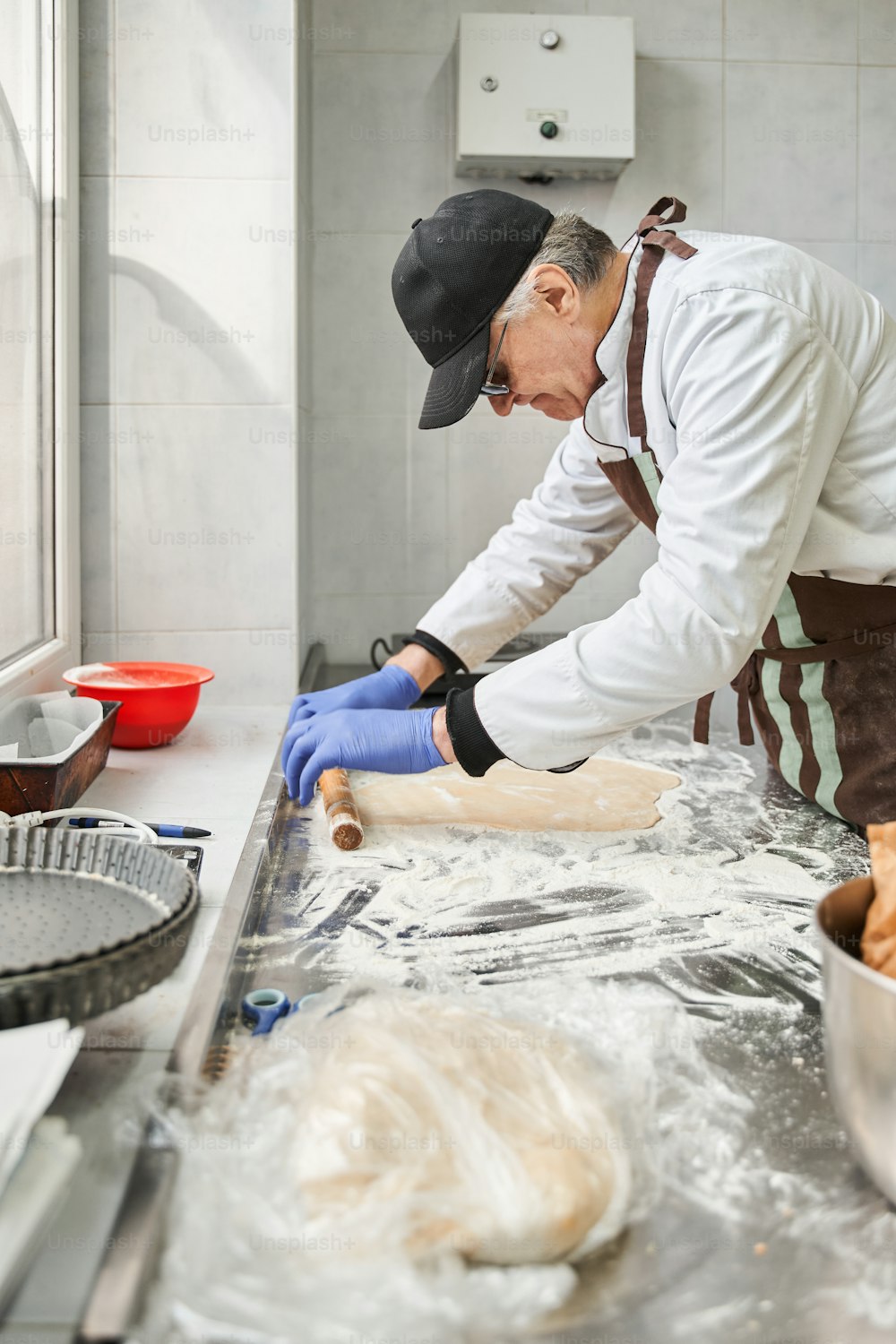 厨房で作業しながら、小麦粉と麺棒でテーブルの上で生地をこねるパン屋の男性の垂直図。仕事のコンセプトでパン屋さんで料理。ストックフォト