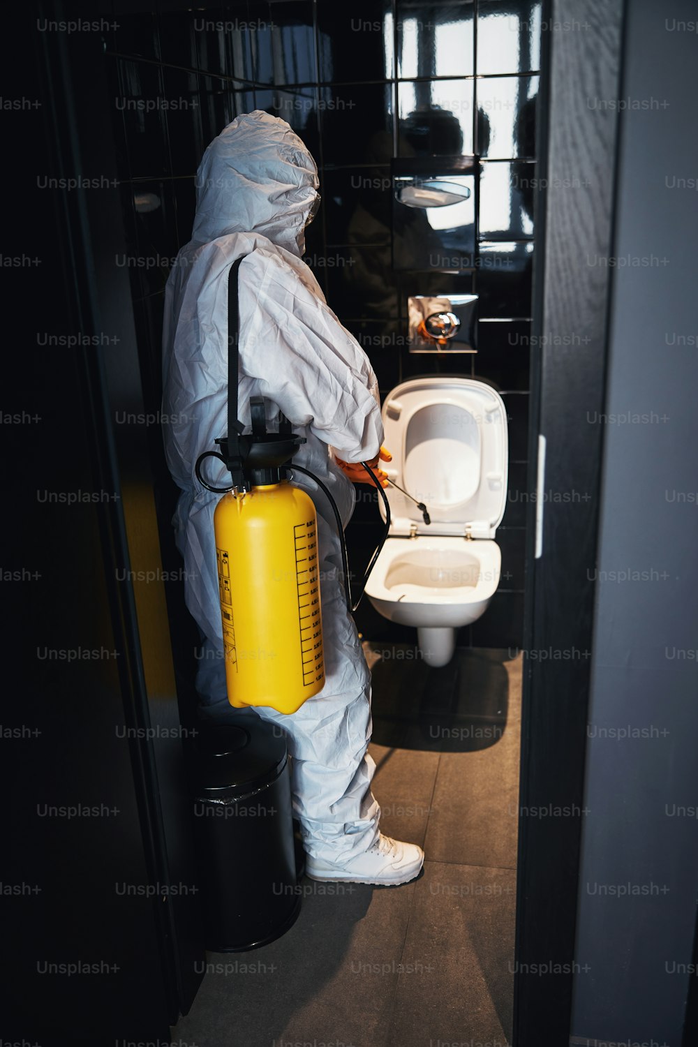 Vue de dos d’un nettoyeur professionnel vêtu d’une combinaison de protection contre les matières dangereuses et de gants en caoutchouc pulvérisant un désinfectant sur la cuvette des toilettes