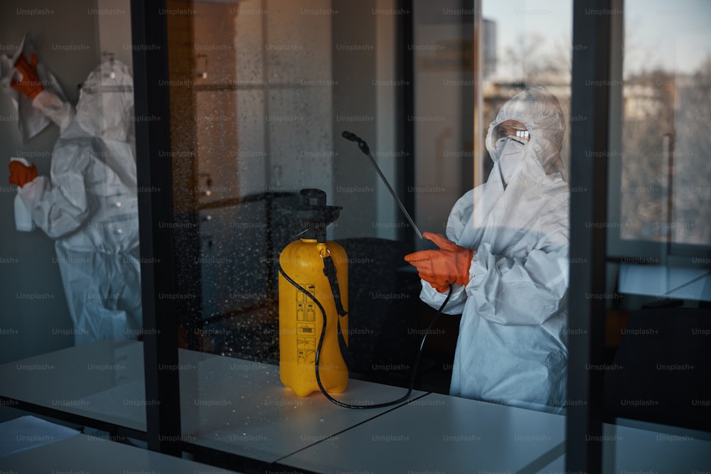 Personnel d’entretien qualifié portant des gants en caoutchouc et des combinaisons de protection contre les matières dangereuses pour désinfecter les surfaces du bureau