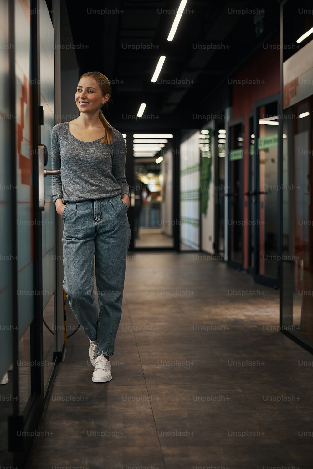 Full-length portrait of a pleased lady with her hands in the trousers pockets standing in the corridor