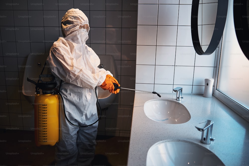 Seitenansicht einer Frau in Schutzbrille und Gummihandschuhen, die das Waschbecken in der öffentlichen Toilette desinfiziert