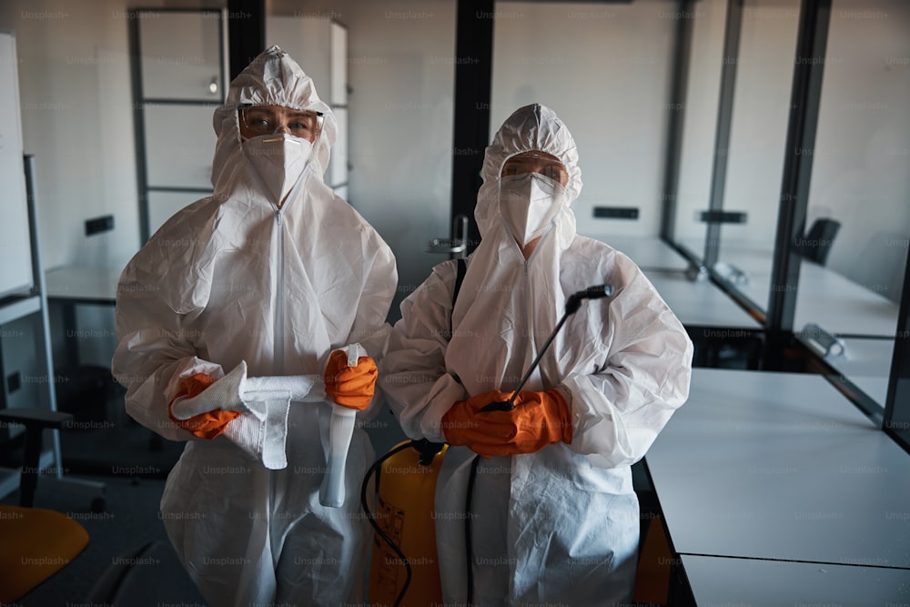 Front view of qualified cleaning staff in rubber gloves and hazmat suits holding janitorial supplies