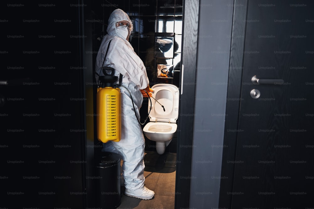 Side view of a trained cleaner in safety goggles and rubber gloves standing by the toilet bowl