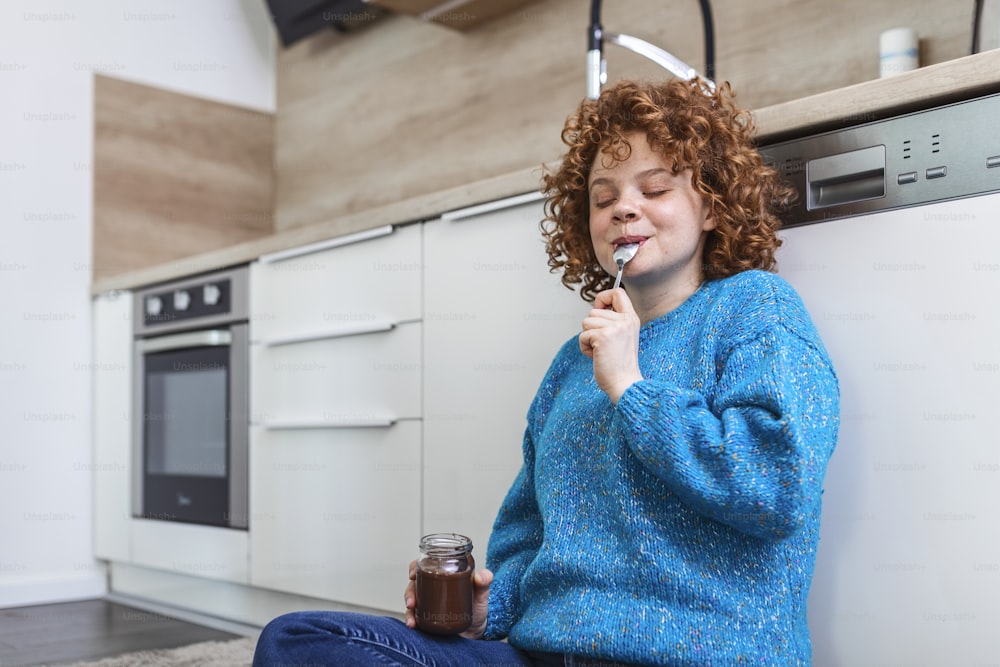 Cute ginger young woman in modern stylish clothes enjoying tasty chocolate spread with cute smile in the cuisine interier. young woman eating chocolate from a jar while sitting on the kitchen floor.