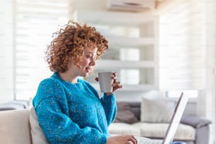 Sorridente jovem ruiva segurando uma xícara de café enquanto trabalhava em seu laptop em sua casa. Menina feliz relaxando no sofá confortável e usando laptop em casa, Internet de navegação