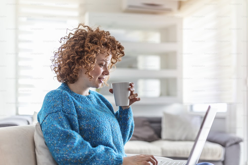 Giovane rossa sorridente che tiene una tazza di caffè mentre lavora sul suo laptop a casa sua. Ragazza felice che si rilassa sul divano comodo e usa il computer portatile a casa, navigando in internet