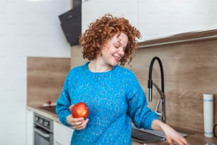 Young beautiful woman with curly red hair eating a juicy red apple while standing in her kitchen at home. Daily intake of vitamins with fruits, Diet and healthy eating
