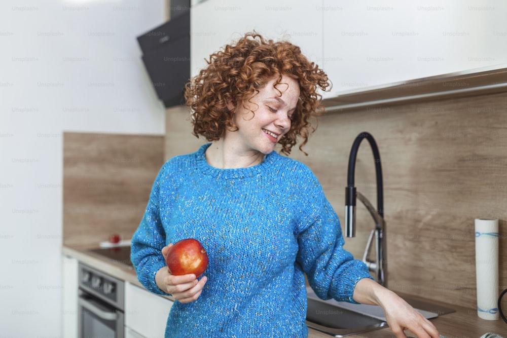 Young beautiful woman with curly red hair eating a juicy red apple while standing in her kitchen at home. Daily intake of vitamins with fruits, Diet and healthy eating
