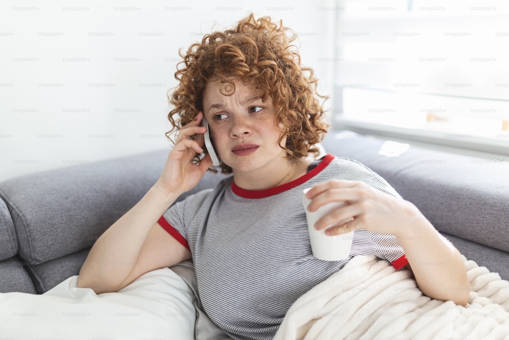 Young woman getting bad news by phone. unhappy woman talking on mobile phone looking down. Crying depressed girl holds phone sitting on sofa hopeless , breaking up, scared of threatening, mobile abuse