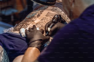 Mãos de um tatuador usando luvas de proteção pretas criando uma imagem nas costas de um homem enquanto segurava uma máquina