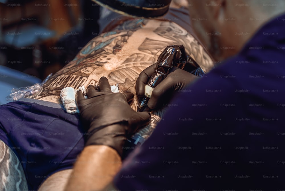Hände eines Tätowierers mit schwarzen Schutzhandschuhen, die ein Bild auf dem Rücken eines Mannes machen, während er eine Maschine hält