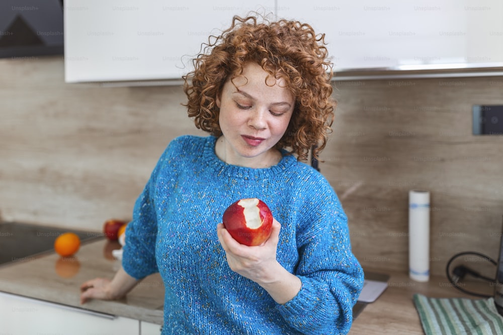Portrait of a lovely smiling pretty girl biting an apple in her kitchen. Young woman enjoying her red organic apple fruit. Daily intake of vitamins with fruits, Diet and healthy eating