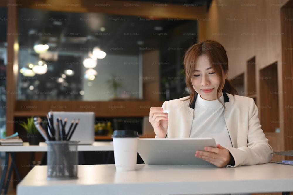 Cadre féminin souriant assis dans un lieu de travail moderne et utilisant une tablette numérique.