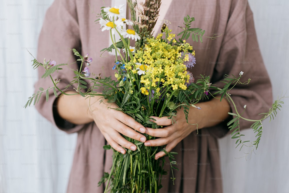 Stylish woman in linen dress holding wildflowers bouquet on background of pastel fabric. Simple slow living. Young female in boho rustic dress with white and yellow meadow flowers in hands