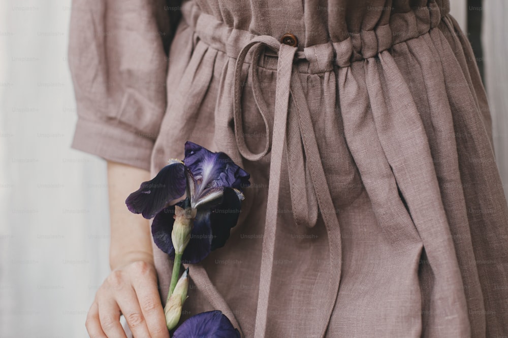 Stylish woman in linen dress holding purple iris flower in sunny light, detail close up. Slow life. Young female in boho rustic dress with iris. Simple aesthetic moment