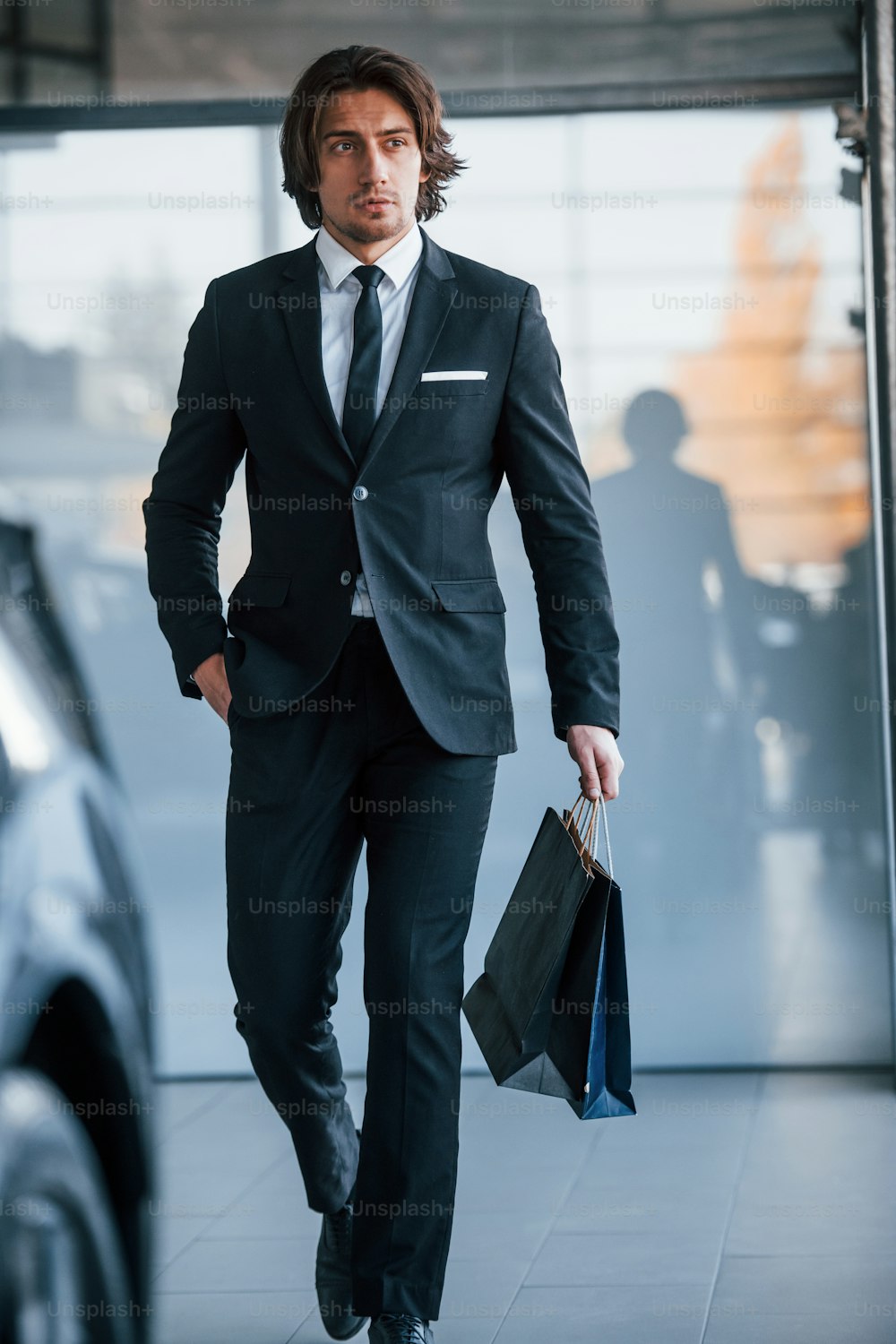 Portrait of handsome young businessman in black suit and tie and with shopping bag near the modern car.
