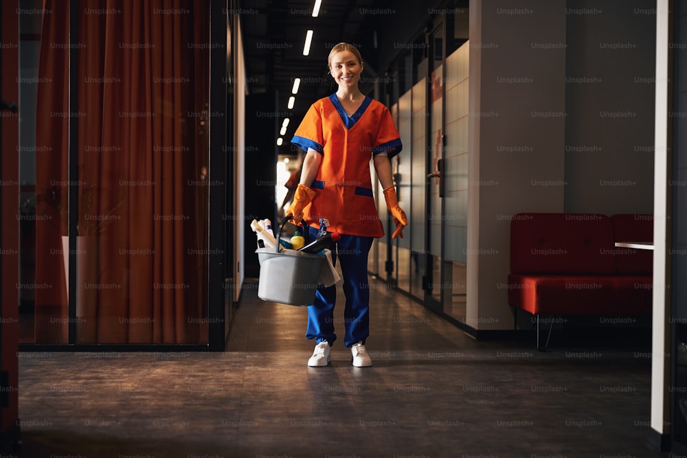Full-length portrait of a joyous young office janitor holding a plastic bucket with cleaning supplies