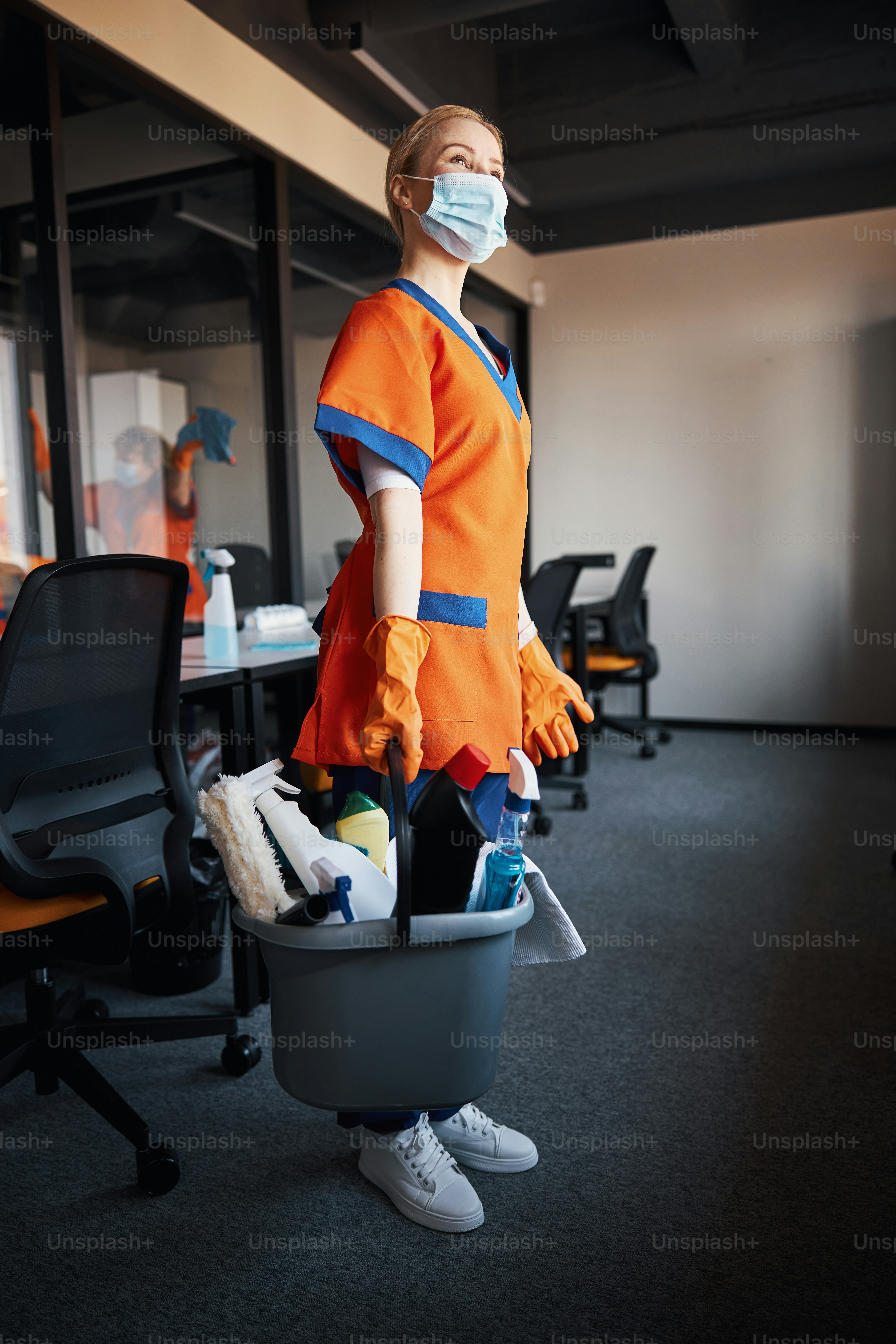 Low angle of a woman in rubber gloves and a face mask holding a plastic bucket with janitorial supplies