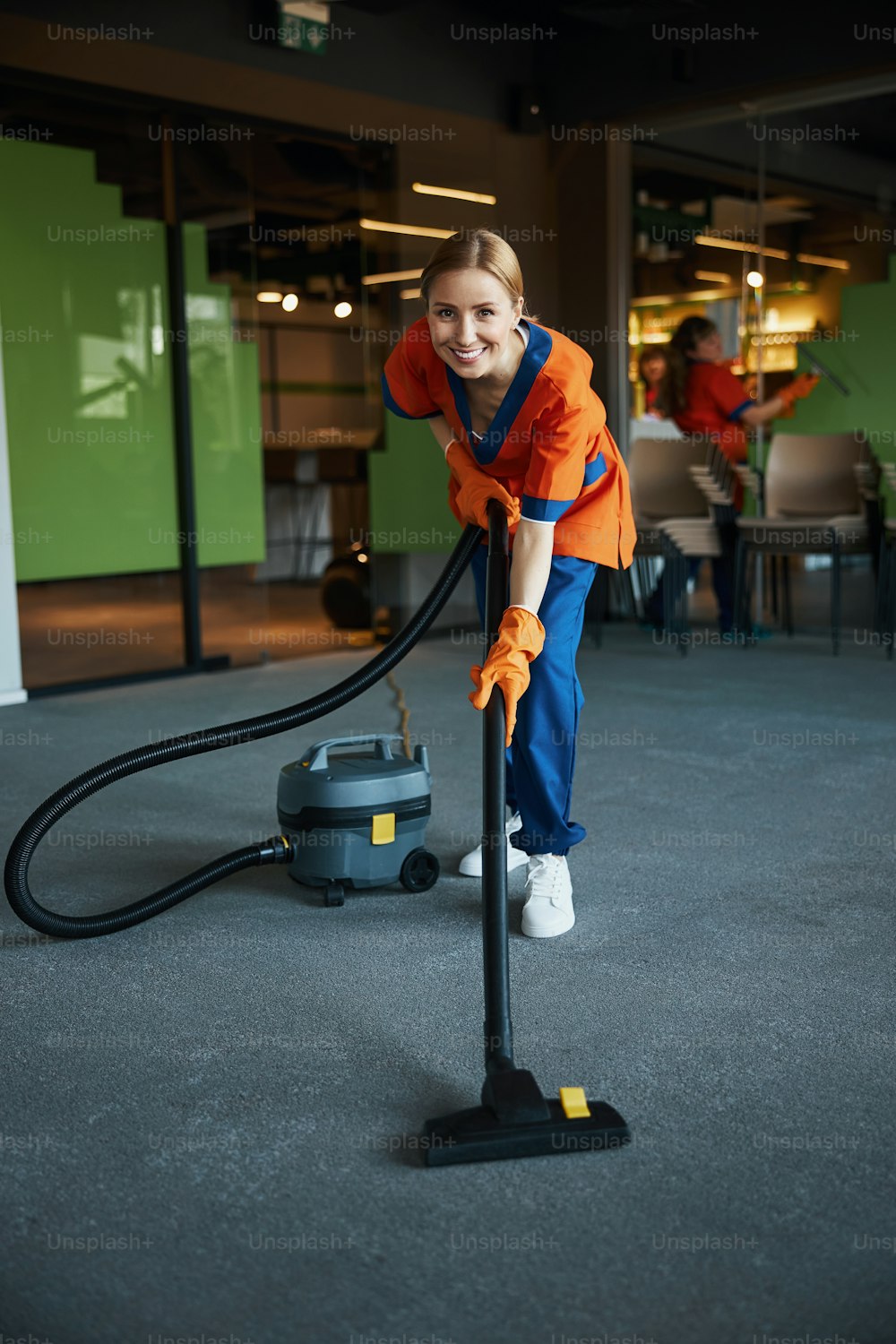 Joyous female worker in the uniform using the corded bagless canister vacuum cleaner during the clean-up