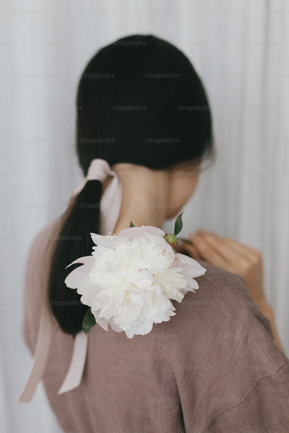 Stylish woman in linen dress holding peony on background of pastel fabric. Slow life. Young female in boho rustic dress hiding face with white peony flower. Simple aesthetic moment