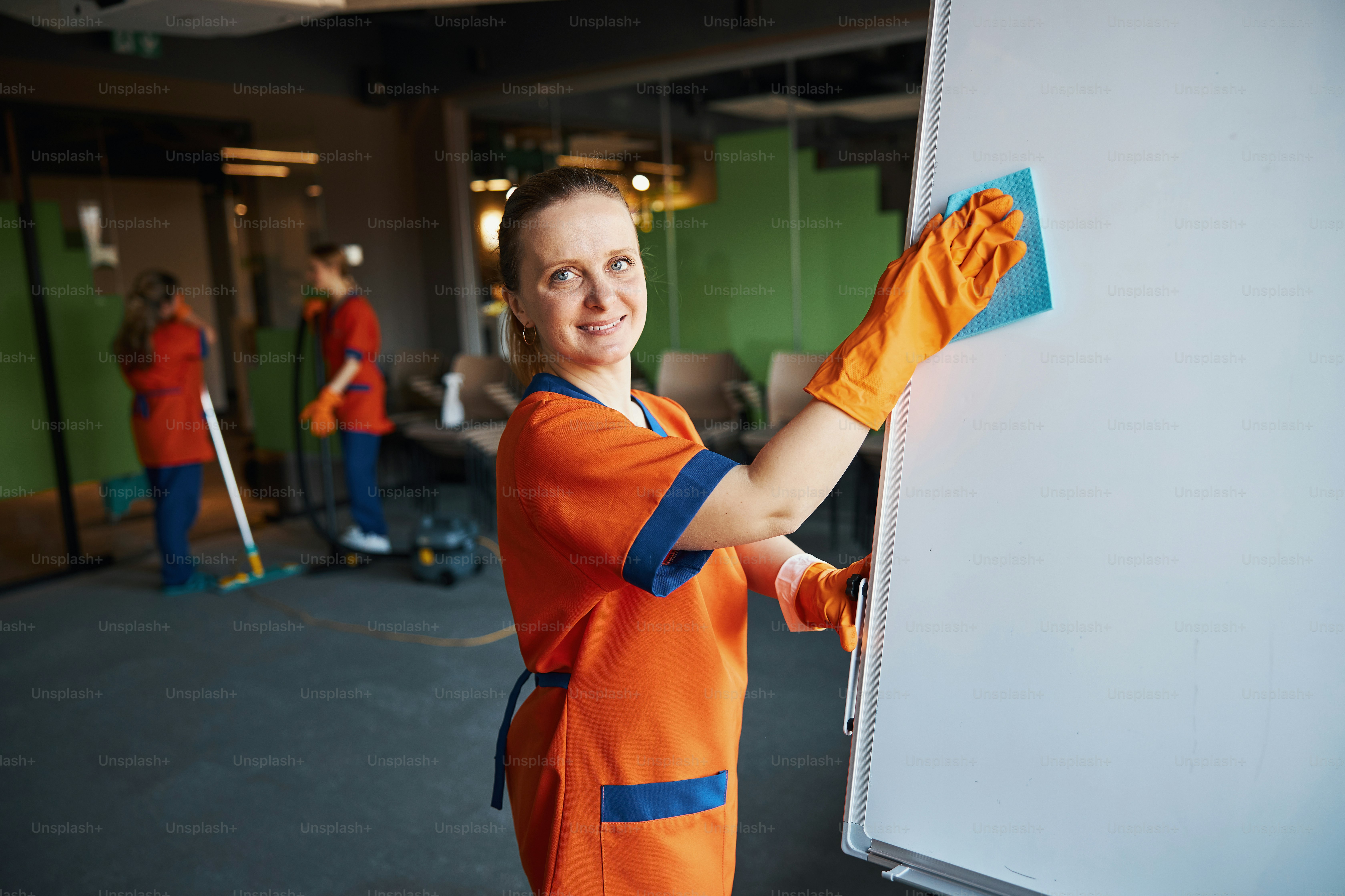 Smiling contented female cleaner wiping down the whiteboard with a wet absorbent cloth during the clean-up