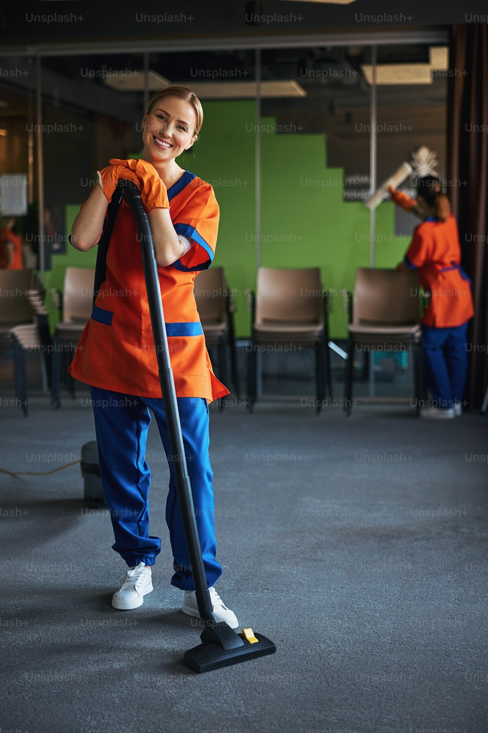 텔레스코픽 진공 청소기 지팡이에 손을 기대고 있는 쾌활한 노동자의 전체 길이 초상화