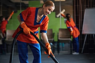 Hermosa trabajadora sonriente y sus colegas usando una aspiradora y escobillas de goma durante la limpieza