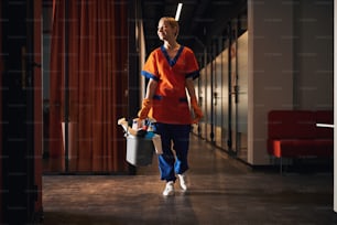 Smiling attractive Caucasian female janitor in rubber gloves carrying a plastic bucket with cleaning products
