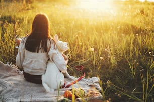 Donna elegante che si gode il tramonto con cane bianco su coperta in una calda luce soleggiata nel prato estivo. Vacanza e picnic con animale domestico. Giovane femmina boho che si rilassa con il cucciolo del pastore svizzero