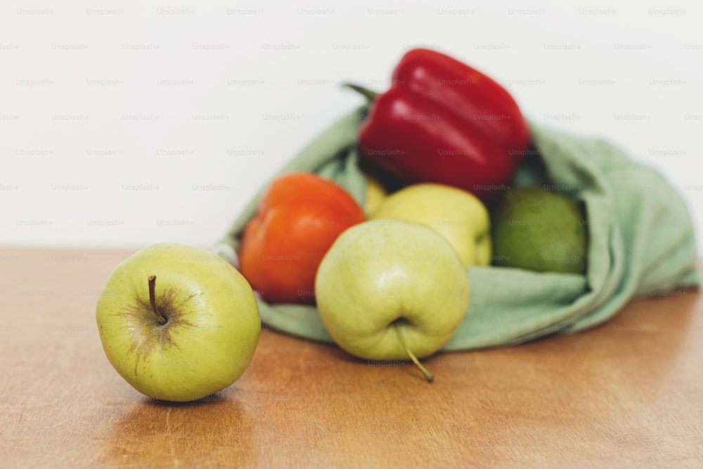 Zero Waste Shopping. Frische Äpfel, Avocado, Tomaten, Paprika im Baumwollbeutel auf Holztisch. Bio-Obst und -Gemüse in wiederverwendbarer Tasche. Umweltfreundliche plastikfreie Lebensmittellieferung