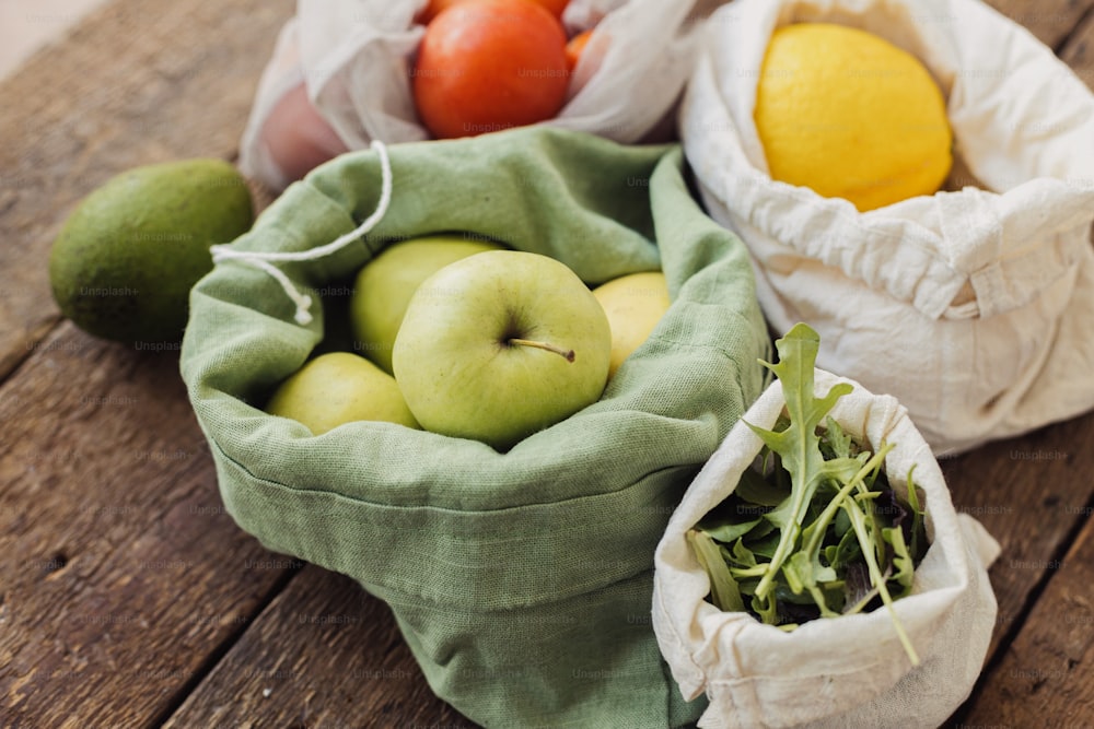 プラスチックフリーの環境に優しい食料品の配達。素朴な木製のテーブルの上に、新鮮なリンゴ、トマト、レモン、アボカド、ルッコラがエココットンの袋に入れられています。廃棄物ゼロ。オーガニックの果物と野菜を再利用可能な袋に入れたもの