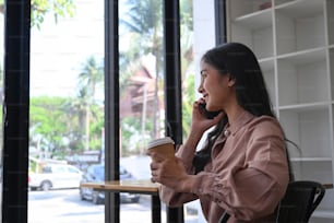 Happy young businesswoman holding coffee cup and talking on mobile phone while sitting near window in office building.