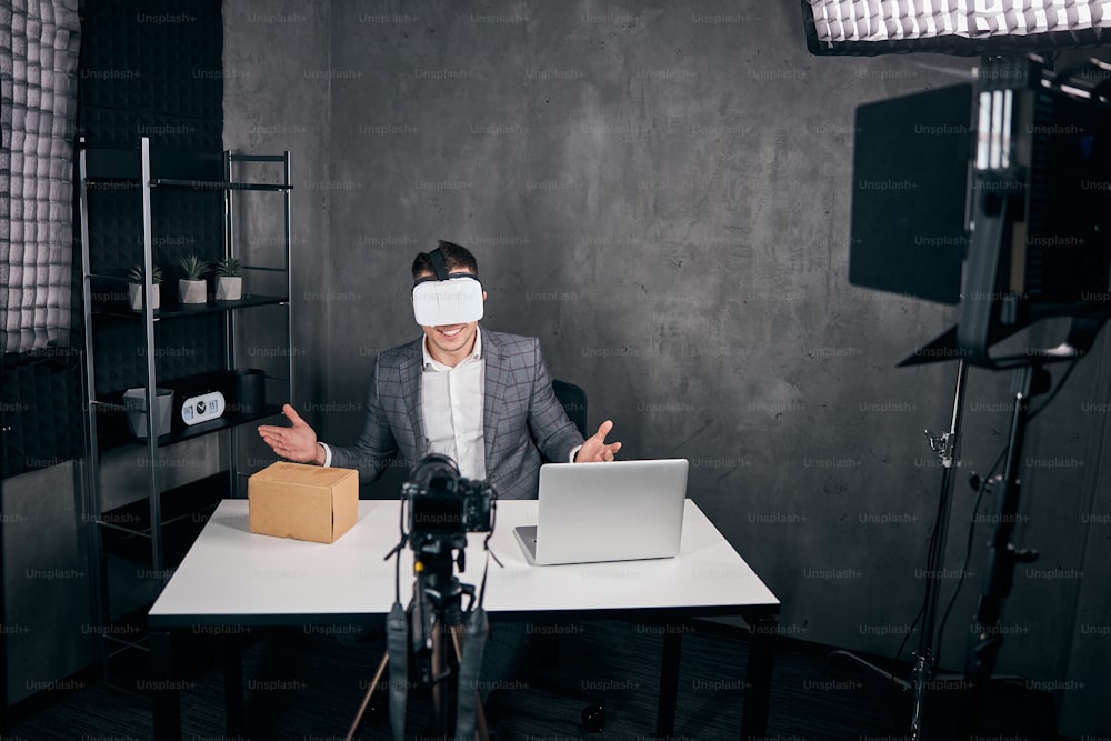 Alegre bloguero masculino que usa gafas de realidad virtual mientras está sentado en la mesa con una computadora portátil y graba video