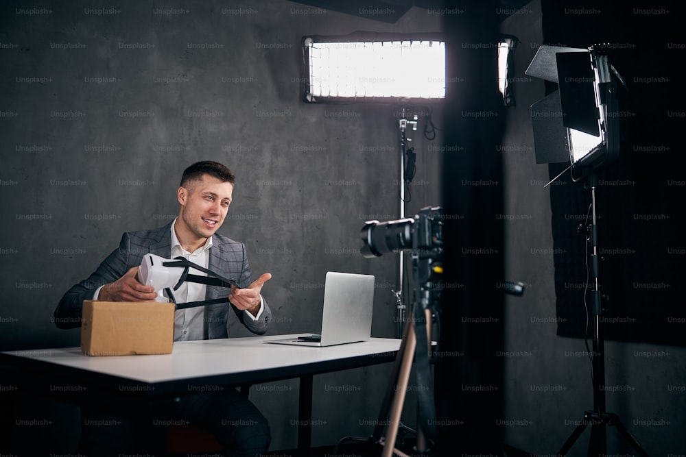 Guapo bloguero masculino sosteniendo gafas de realidad virtual y sonriendo mientras está sentado en la mesa con una computadora portátil y filmando videos para vlog