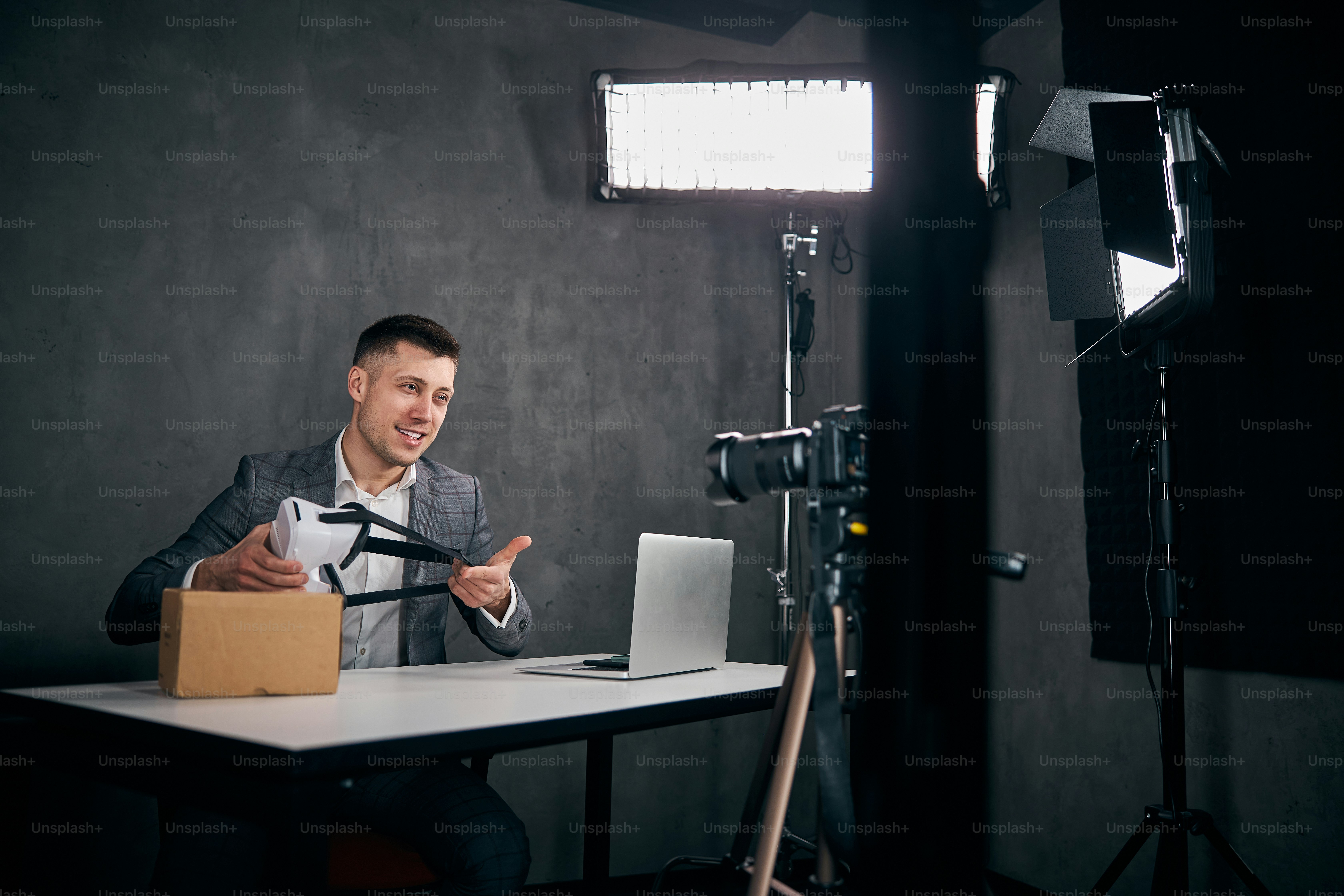 Handsome male blogger holding VR glasses and smiling while sitting at the table with laptop and filming video for vlog