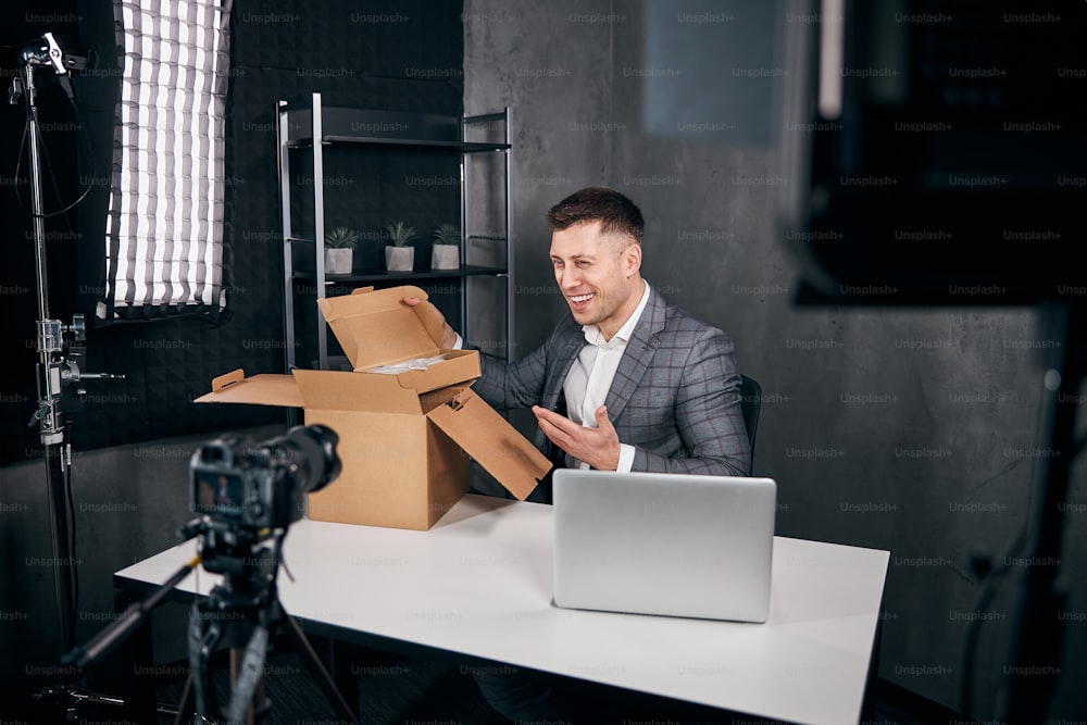 Blogueiro masculino alegre desembalando caixa com compras e sorrindo enquanto filma vídeo para o blog