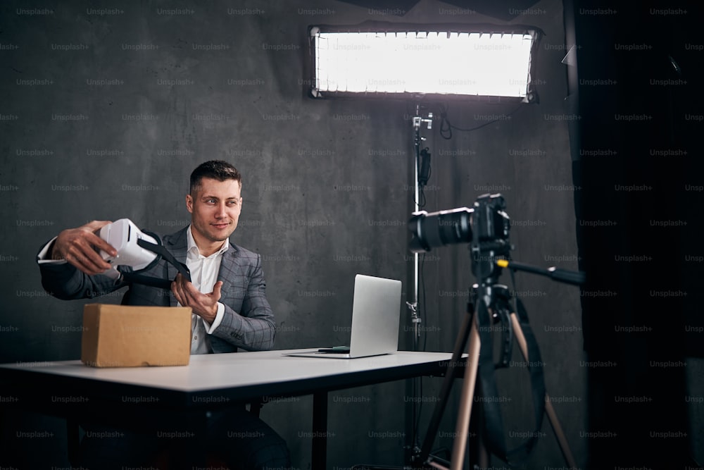 Blogueiro masculino alegre segurando óculos VR e sorrindo enquanto se senta à mesa com laptop e filmando vídeo com câmera profissional
