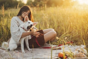 Stylish happy woman playing with her white dog with herb on blanket in warm sunny light in summer meadow. Summer vacation and picnic with pet. Young boho female relaxing with swiss shepherd puppy