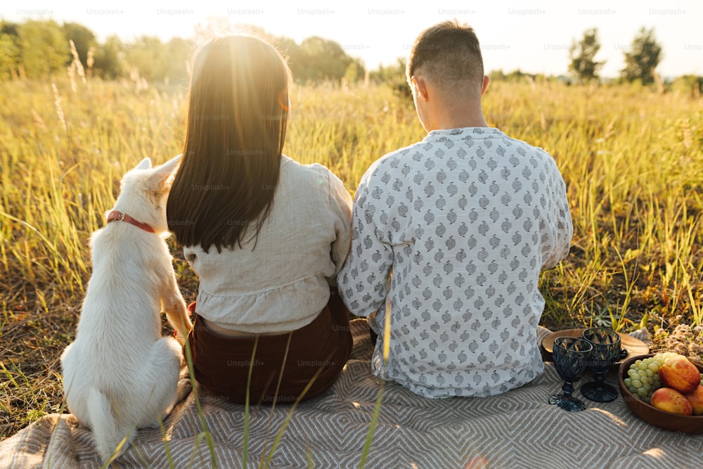 Stylish beautiful couple with white dog relaxing on blanket in warm sunny light among grass in summer meadow. Summer vacation and picnic. Young family enjoying sunset with swiss shepherd puppy