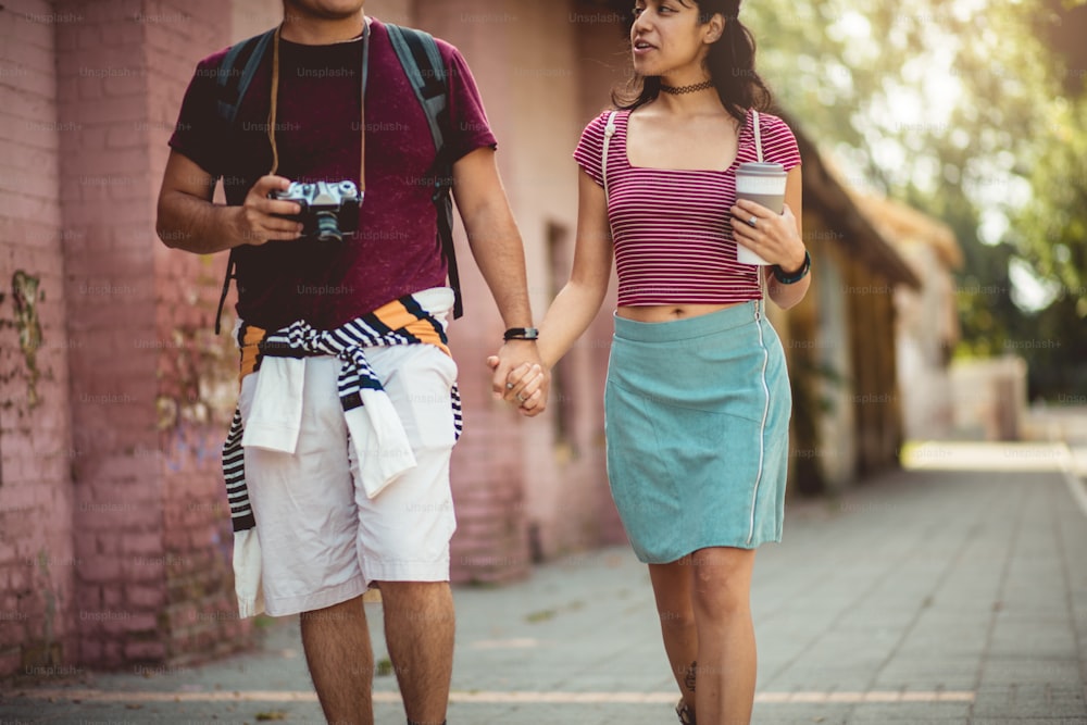 Junges Paar, das durch die Straße geht und Händchen hält.  Mann mit Kamera und Mädchen mit Tasse Kaffee.