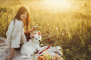Stylish happy woman relaxing with her white dog on blanket in warm sunny light in summer meadow. Vacation and picnic with pet. Young boho female enjoying sunset with swiss shepherd puppy