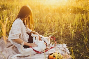 Stylish happy woman playing with her white dog on blanket in warm sunny light in summer meadow. Summer vacation and picnic with pet. Young boho female relaxing with swiss shepherd puppy in sunset