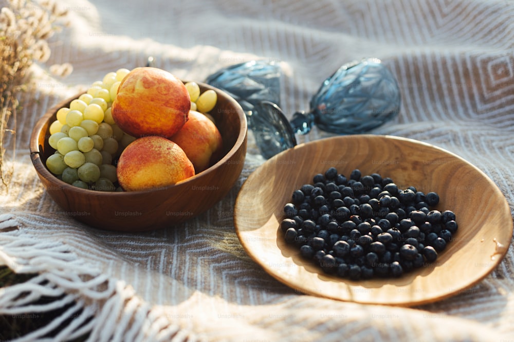 Deliciosas frutas frescas en platos de madera y copas de vino azules sobre cuadros amarillos a la luz cálida y soleada en el prado de verano. Concepto de picnic de verano. Vacaciones y ocio